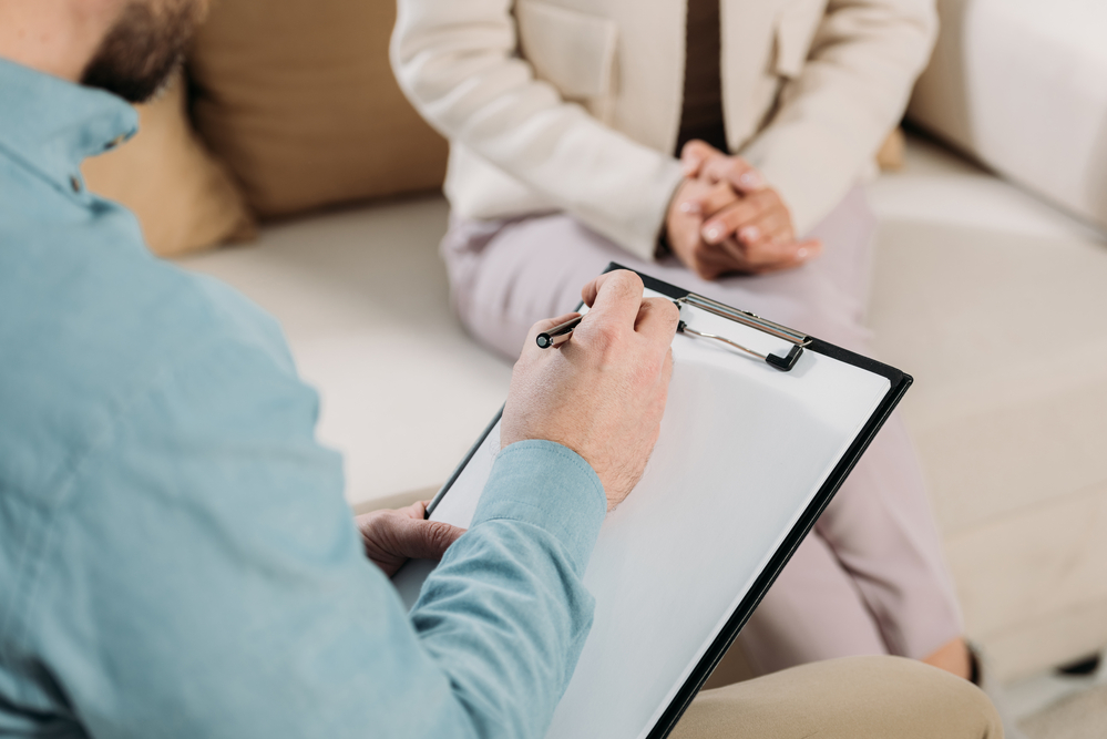 Cropped shot of Psychiatrist writing on clipboard and female patient sitting on couch concept image for mental health practice.